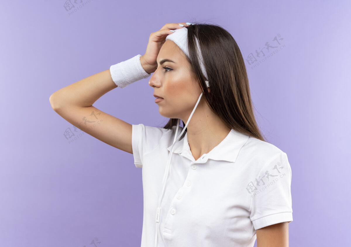侧面漂亮的运动女孩戴着头带和腕带 戴着耳机 手放在头上 看着紫色空间里孤立的一面手放好看