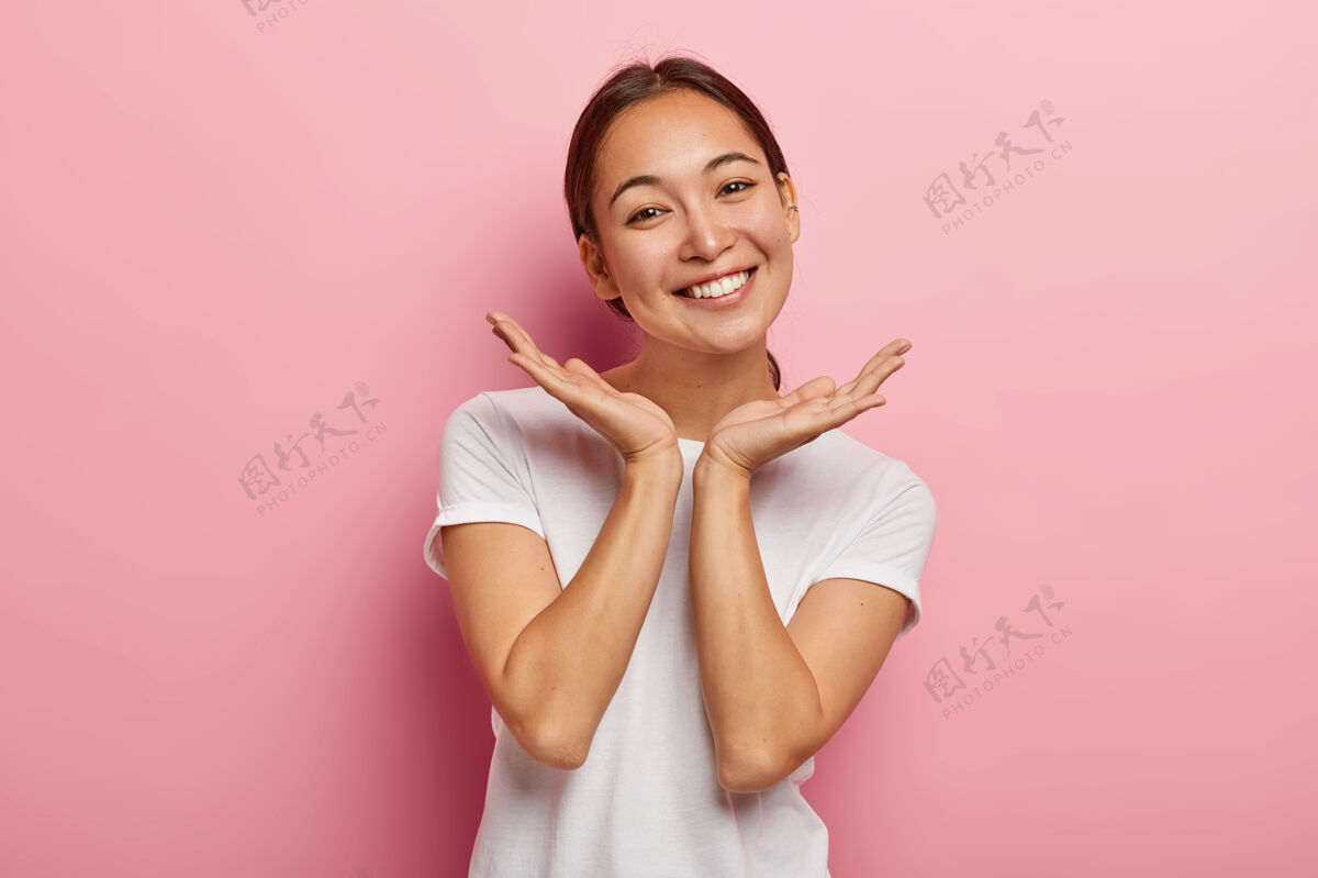 特写容貌宜人的亚洲女模特笑容可掬 掌心贴脸 表达积极的情感 穿着白色t恤 外表迷人 皮肤健康 隔离在粉红色的墙上快乐积极休闲