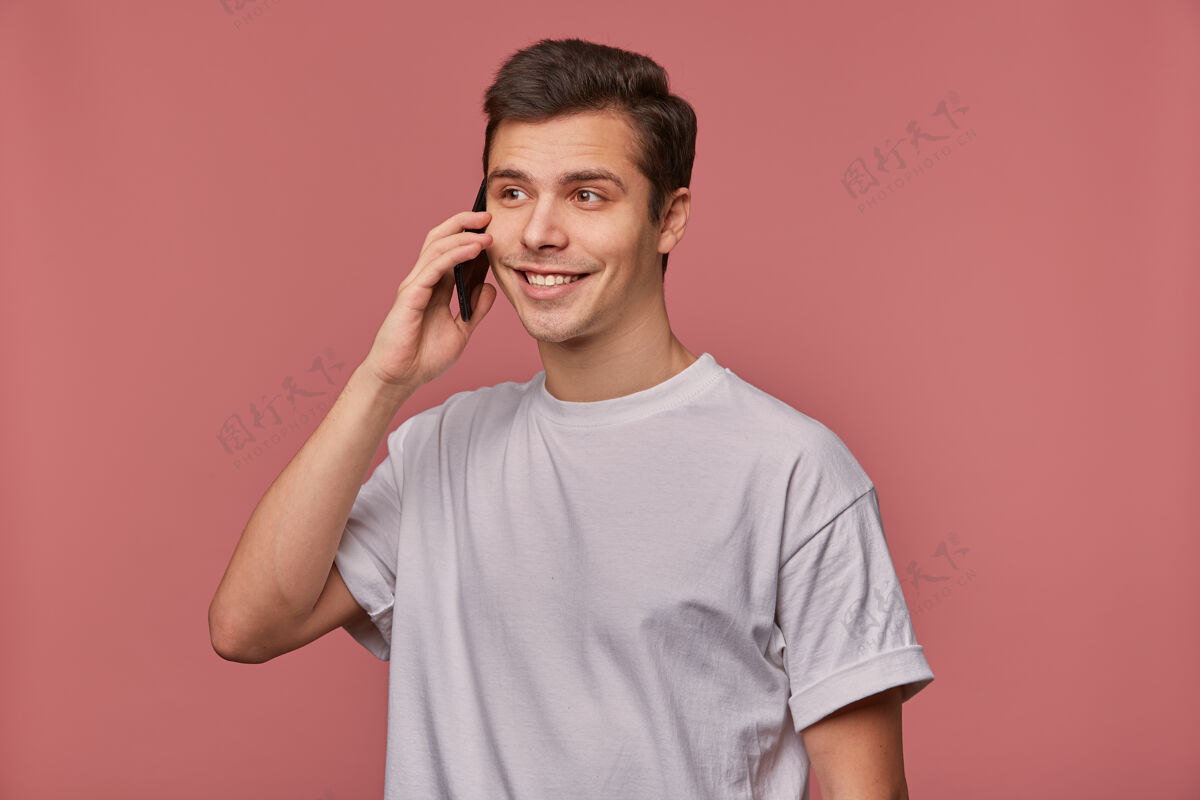 友好一个年轻开朗的人的肖像 穿着一件白t恤 在电话里说话 看起来很高兴 站在粉红色的椅子上积极酷粉色