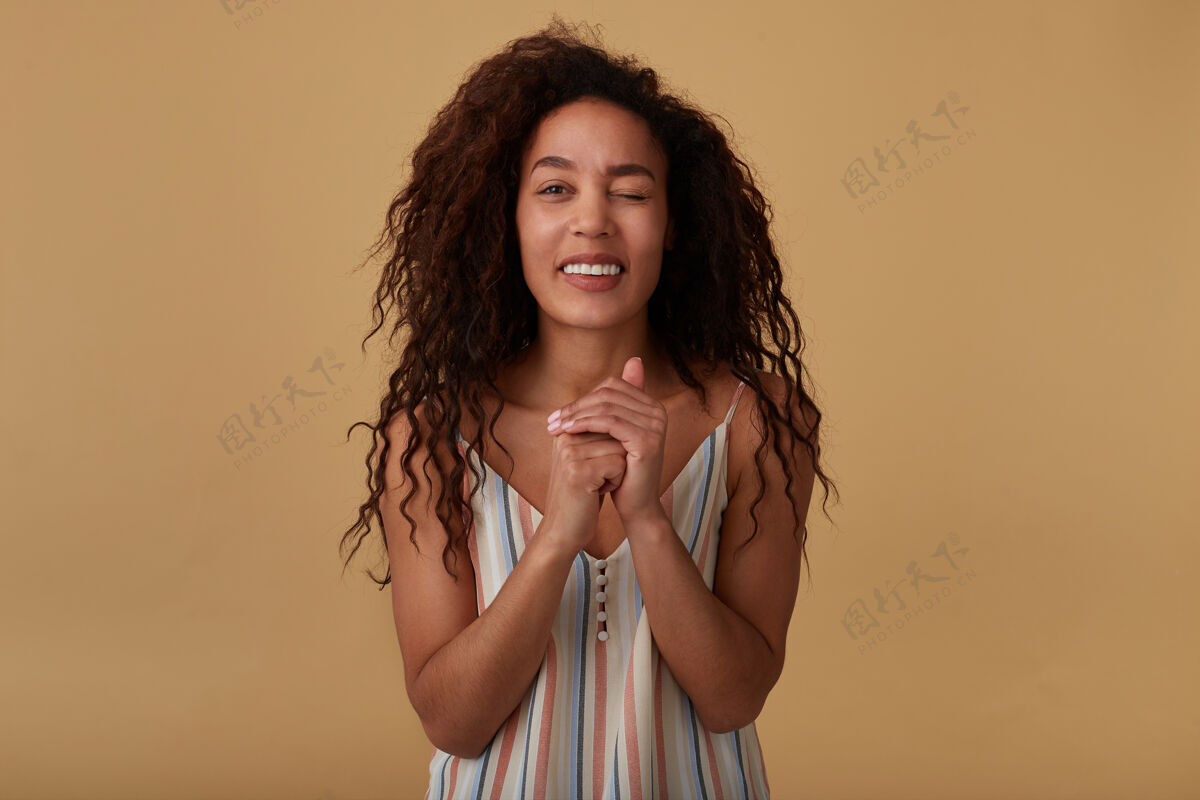 凸起积极的年轻卷曲的黑皮肤黑发女子在夏季条纹衬衫保持举起的双手交叉 并给予耀眼的眨眼 在米色隔离摩卡折叠休闲