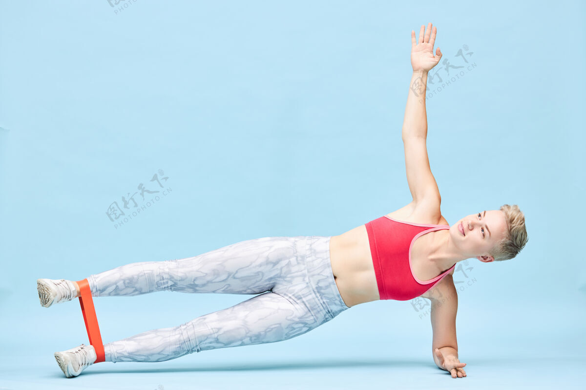 运动一个漂亮的充满活力的运动女孩在健身房的地板上表演侧支撑 脚踝上缠着阻力带做侧举 加强髋外展肌 微笑阻力运动员健身