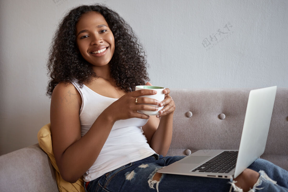 现代美丽迷人的非洲年轻女子在她20多岁的侧视图 大学毕业后在家休息 躺在沙发上 膝上放着便携式电脑 上网 网上购物 面带微笑马克杯茶咖啡