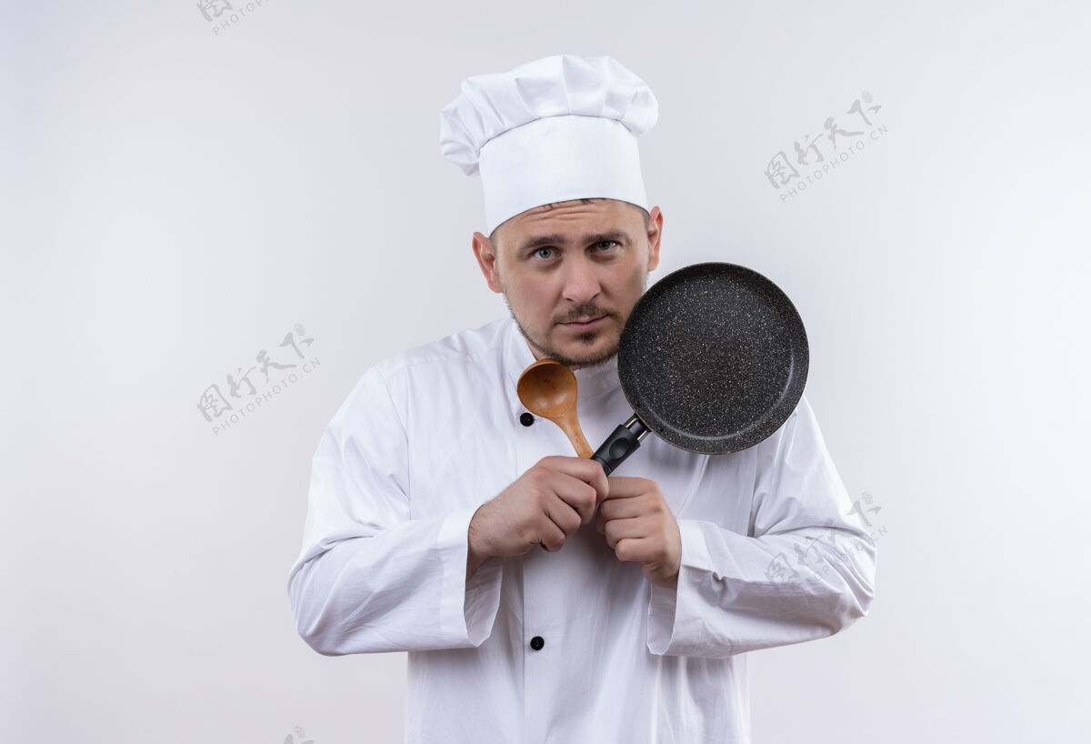 持有体贴的年轻帅哥厨师穿着厨师制服拿着勺子和煎锅隔离在空白处锅厨师薯条
