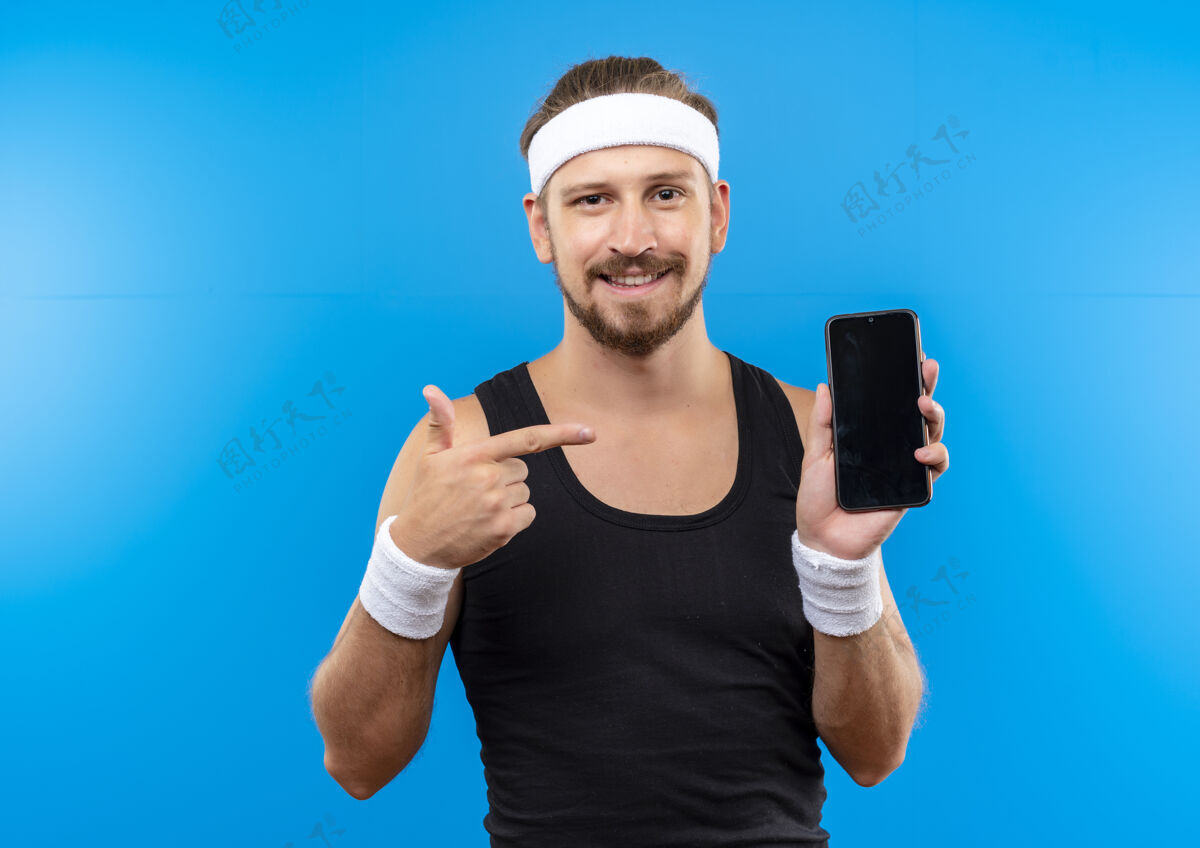 微笑面带微笑的年轻帅气的运动型男人戴着头带和腕带拿着手机指着蓝色的空间手持年轻帅气