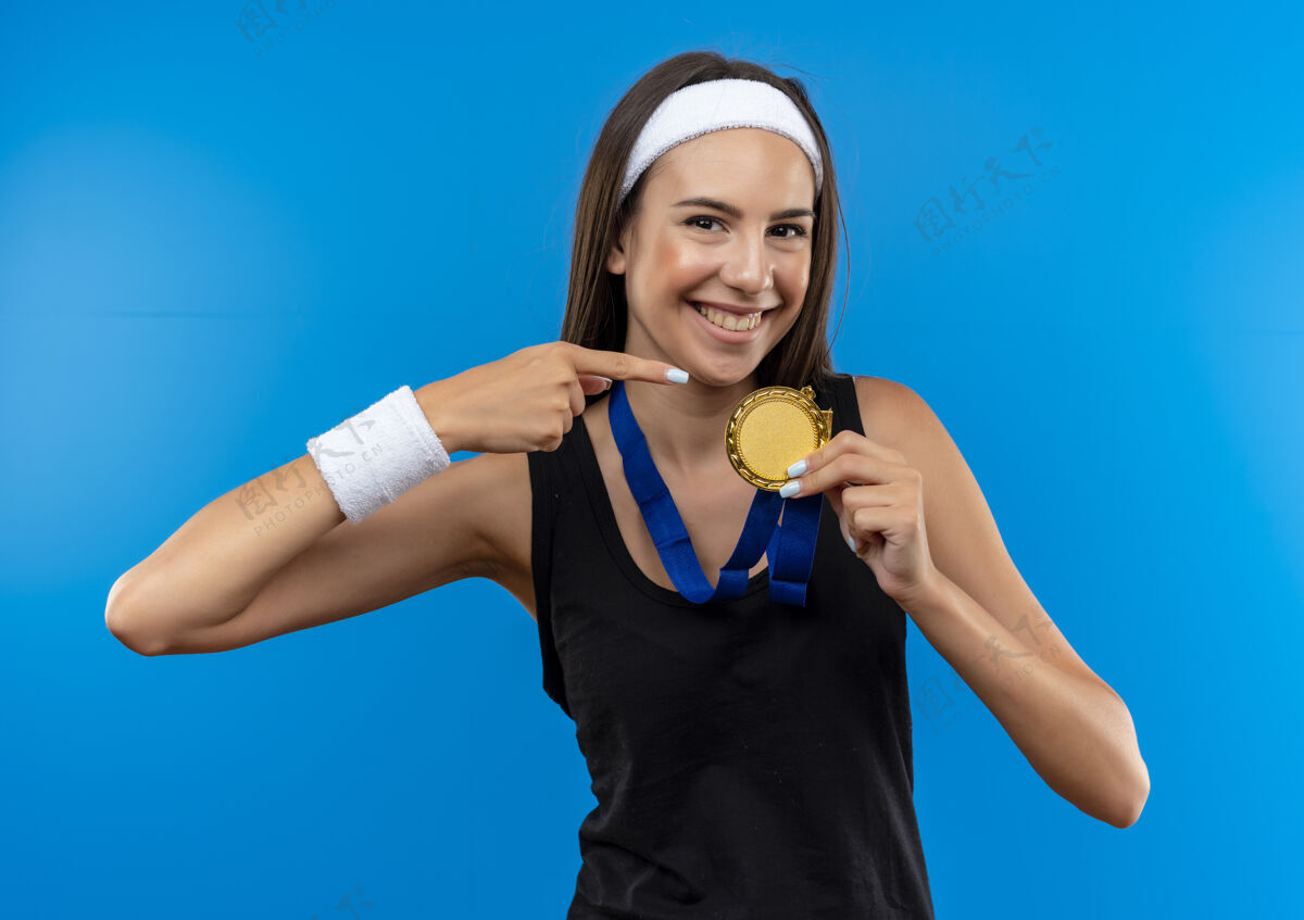 微笑微笑着的年轻漂亮的运动女孩戴着头带和腕带 脖子上戴着勋章 指着孤立在蓝色空间的勋章腕带运动奖牌