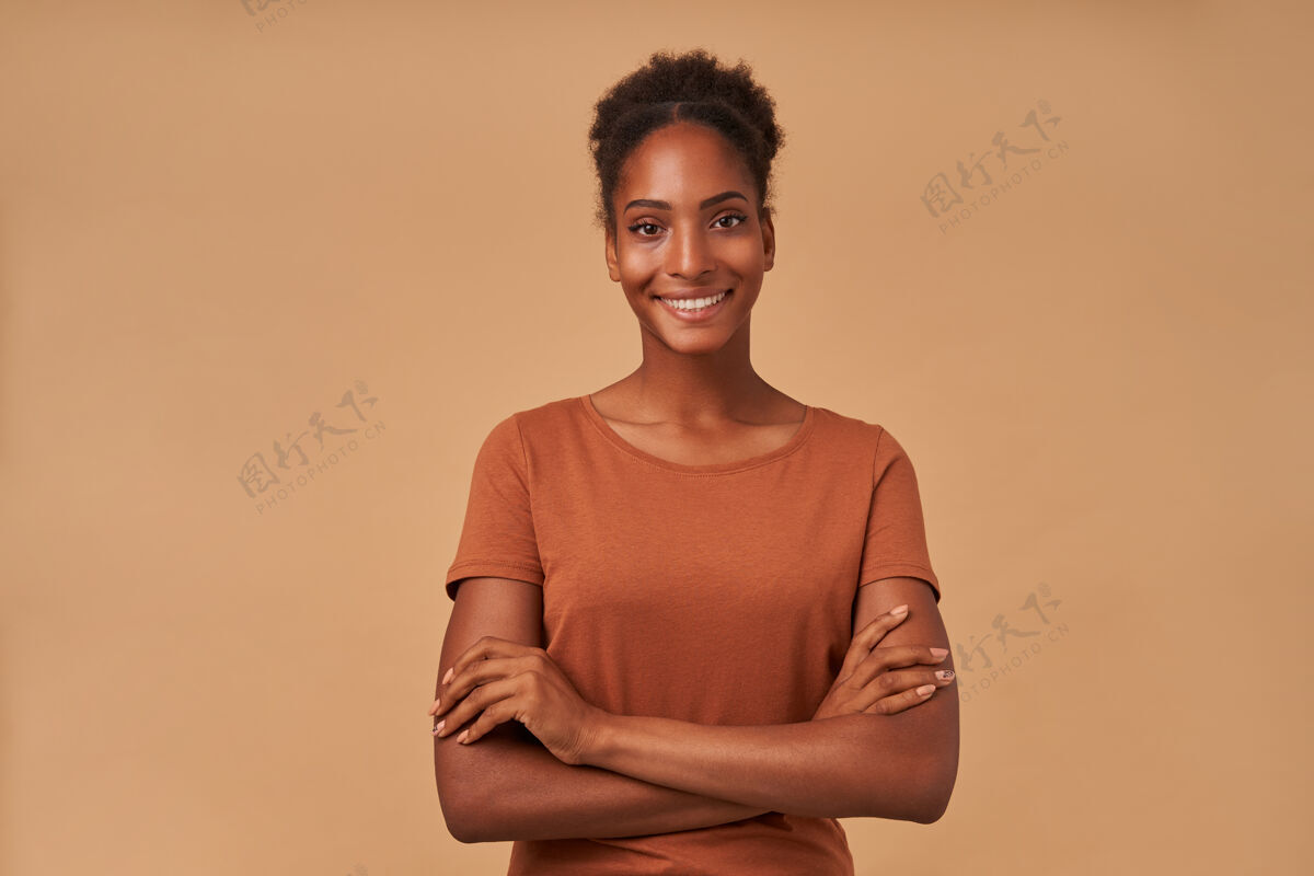 高年轻漂亮的黑皮肤卷发女士的肖像 双手交叉 面带迷人的微笑 站在米色的地面上化妆品非洲皮肤