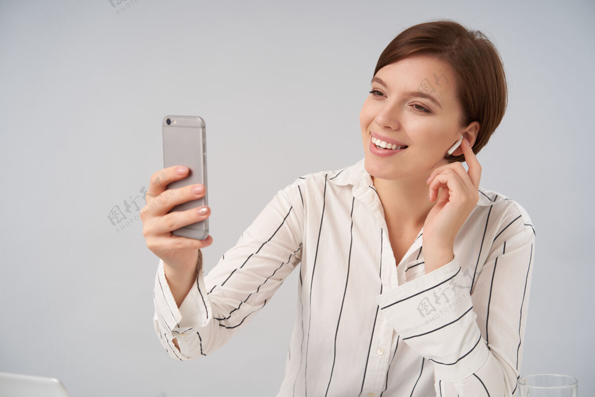 情绪可爱的棕色头发的年轻女性的肖像 短发时髦 微笑积极 同时与手机和耳机视频聊天 隔离在白色短优雅积极