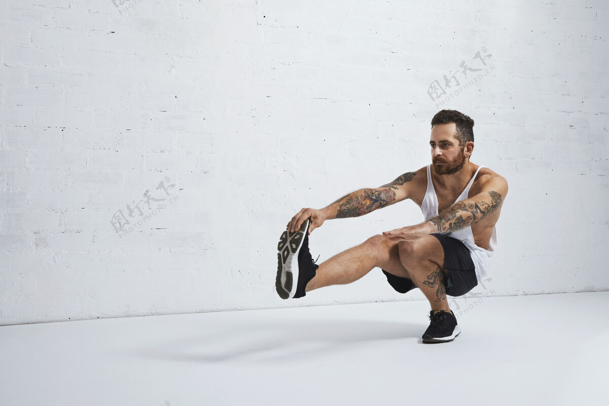 健身残酷的纹身健美操教练秀操动作一条腿蹲下 隔离在白砖墙上训练体重锻炼