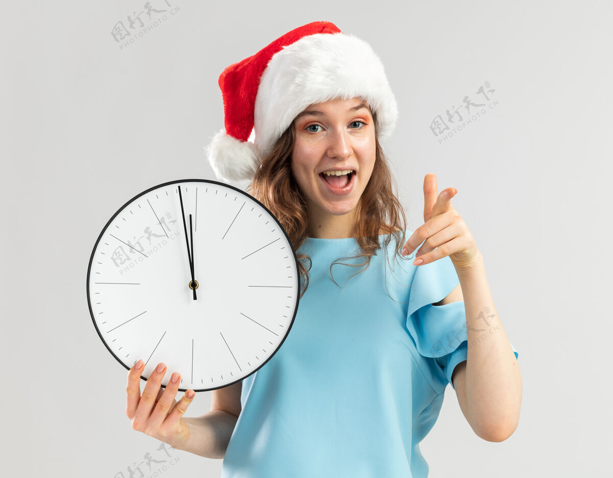 圣诞快乐戴着蓝色上衣和圣诞帽的年轻女子拿着挂钟 用食指指着 快乐而积极手指指数新年