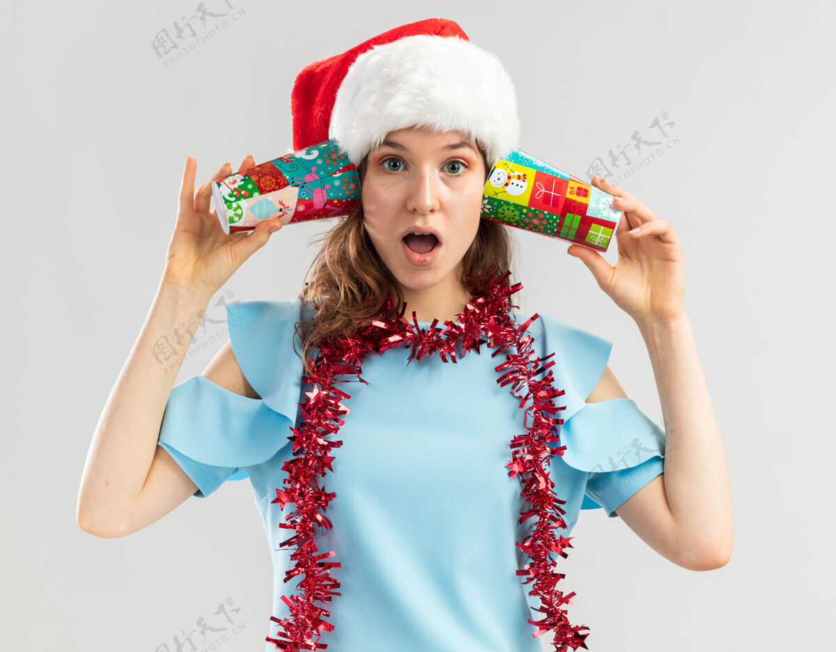 惊讶身穿蓝色上衣 戴着圣诞帽 脖子上戴着金属丝的年轻女子 耳朵上戴着五颜六色的纸杯 看上去很惊讶脖子圣诞快乐帽子