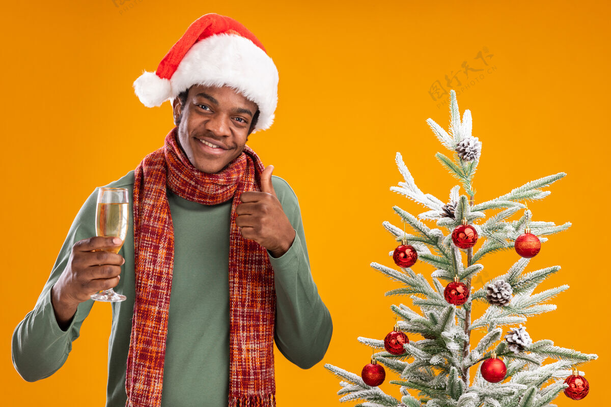 圣诞节戴着圣诞帽 围着围巾的非洲裔美国人 手拿香槟酒杯 在橙色背景下的圣诞树旁愉快地竖起大拇指展示微笑玻璃杯