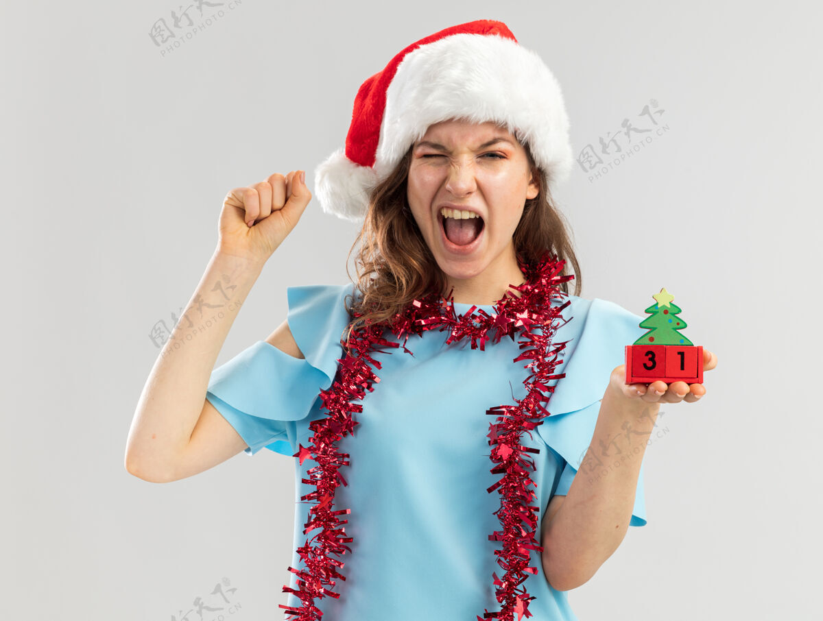 新年穿着蓝色上衣 戴着圣诞帽 脖子上戴着金属丝的年轻女子 手里拿着玩具立方体 手里握着新年快乐的约会对象 握紧拳头尖叫着庆祝疯狂的快乐尖叫顶蓝色