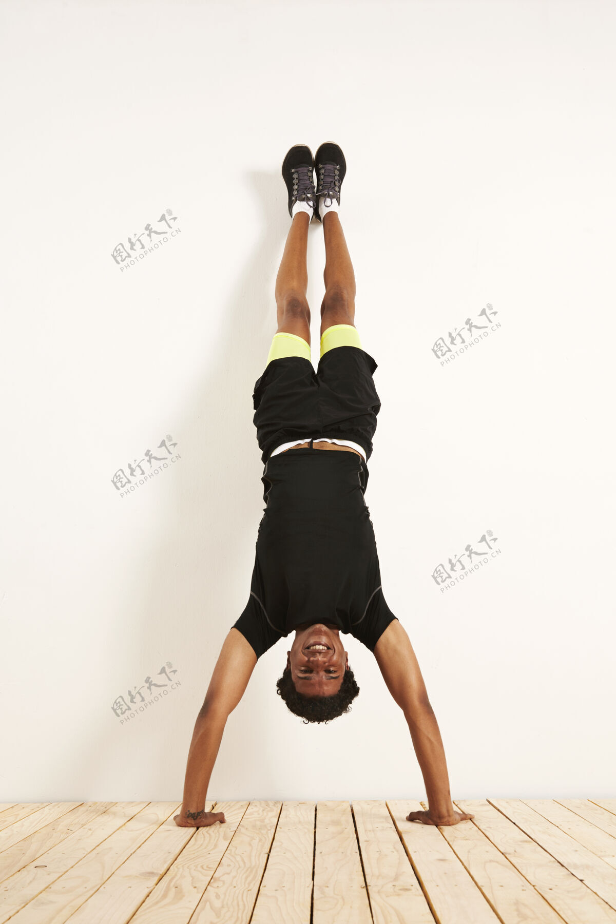 运动快乐微笑的年轻黑人模特穿着黑黄相间的运动服 在木地板上的白墙上做倒立动作肌肉运动员健身