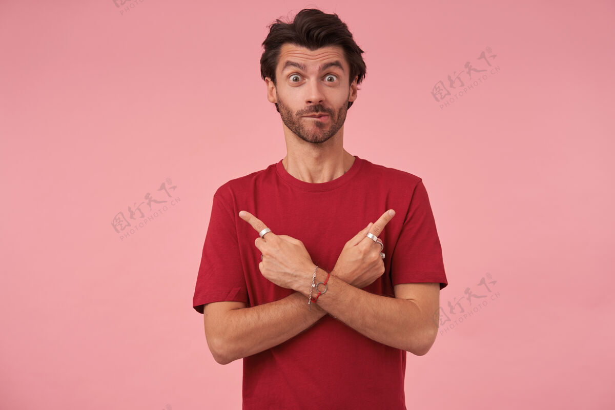 不同惊讶的年轻人 时髦的发型 食指指向不同的方向 站在粉红色的休闲服上扬起眉毛 咬着屁股正面男性男人
