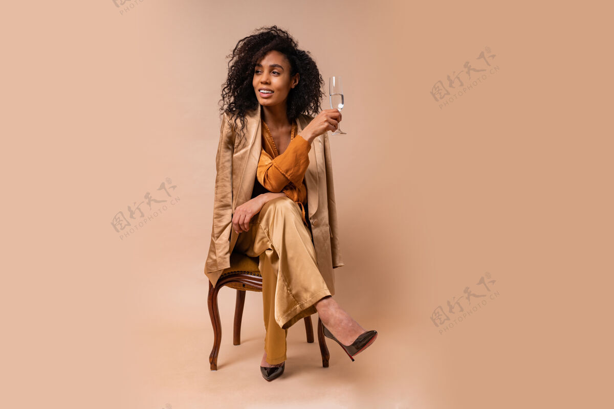 年轻快乐的女人 皮肤黝黑 头发卷曲 手里拿着一杯酒 穿着一件漂亮的橙色衬衫和丝绸裤子 坐在老式椅子米色的墙上奢华前面风景