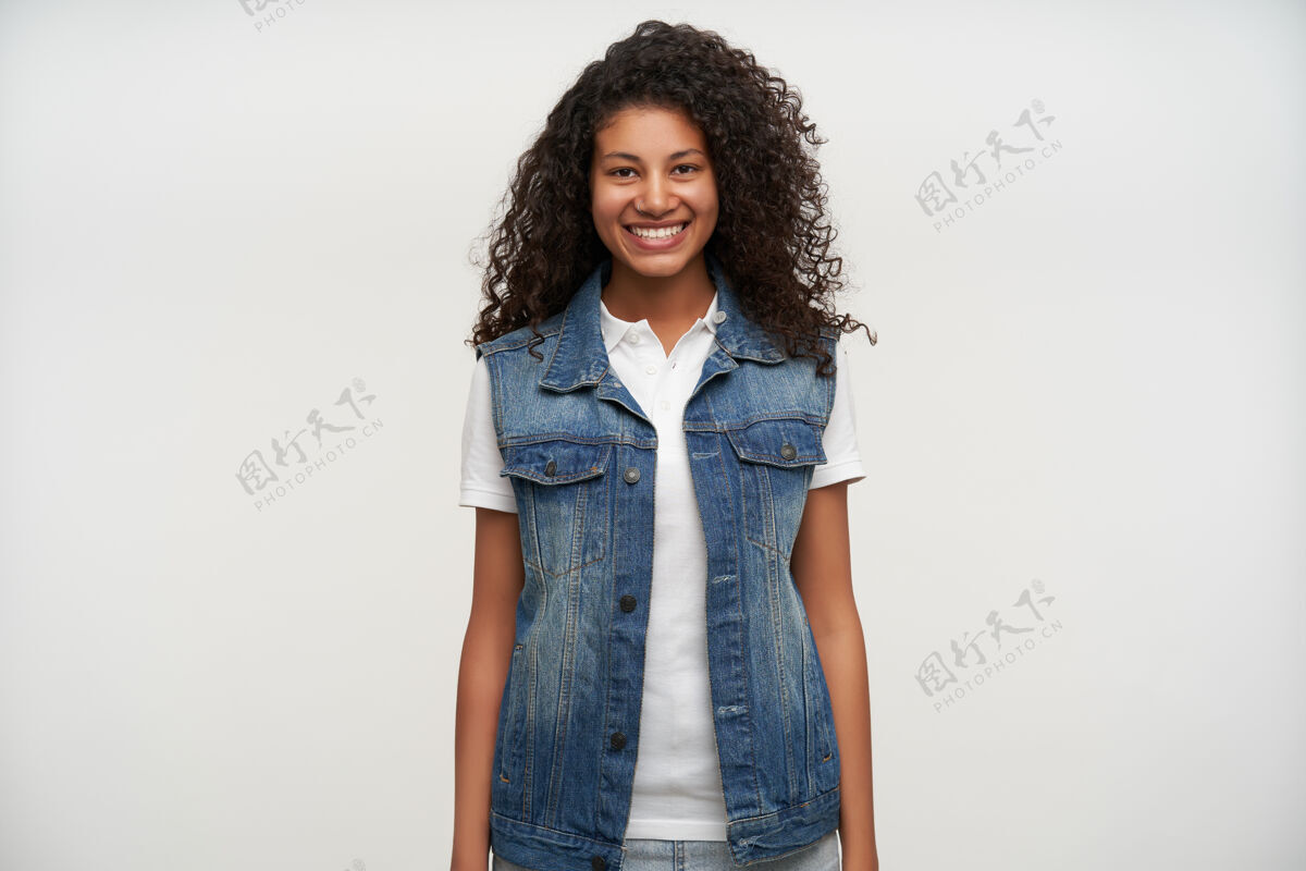 女性一幅年轻的黑皮肤女人的肖像 棕色卷发 双手挽着身体 幸福地微笑着 穿着休闲服在白色的衣服上摆姿势室内皮肤快乐