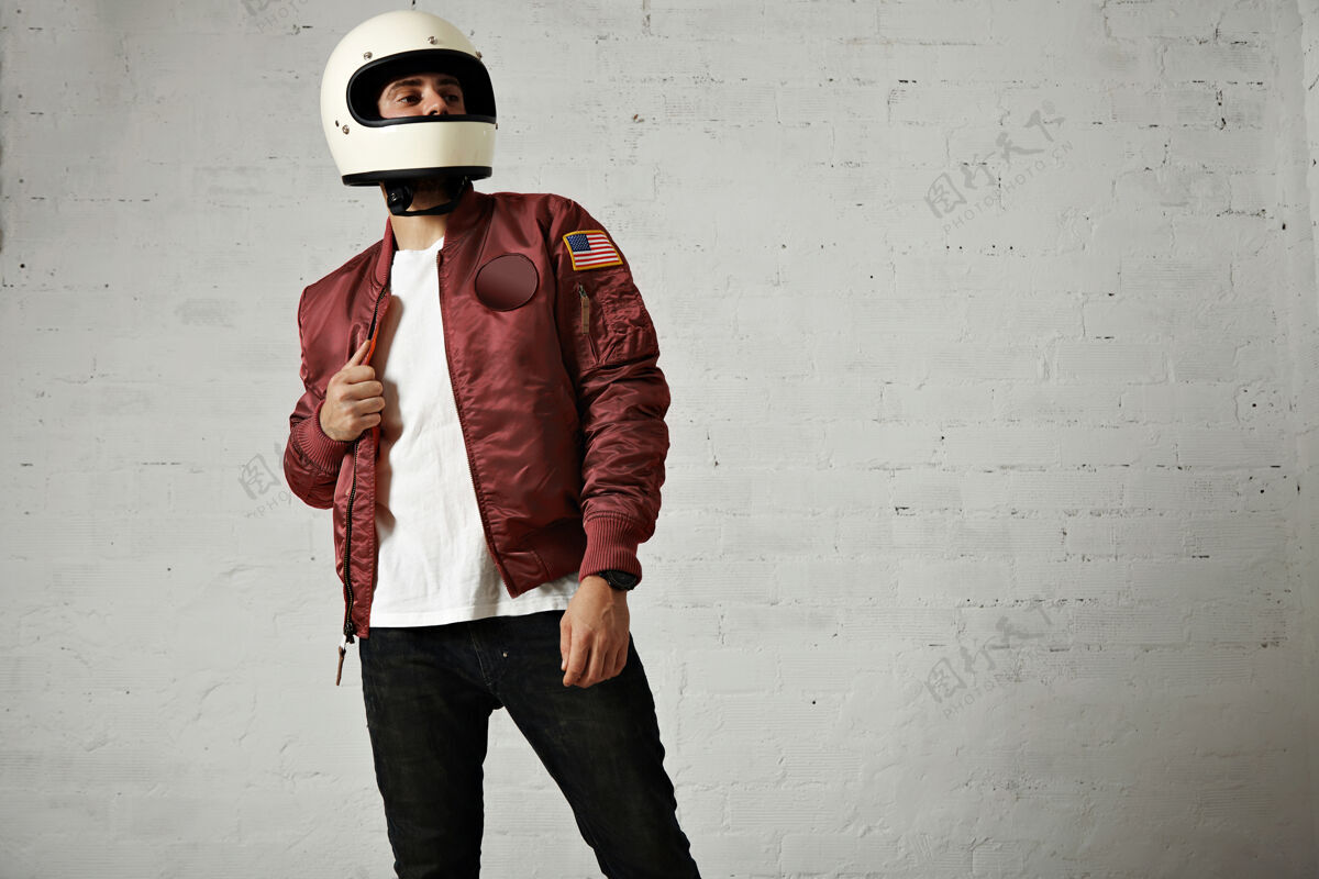 男人骄傲的摩托车手在一个普通的白色头盔 波尔多尼龙棒球服夹克 牛仔裤和白墙背景下的t恤酷城市帽子