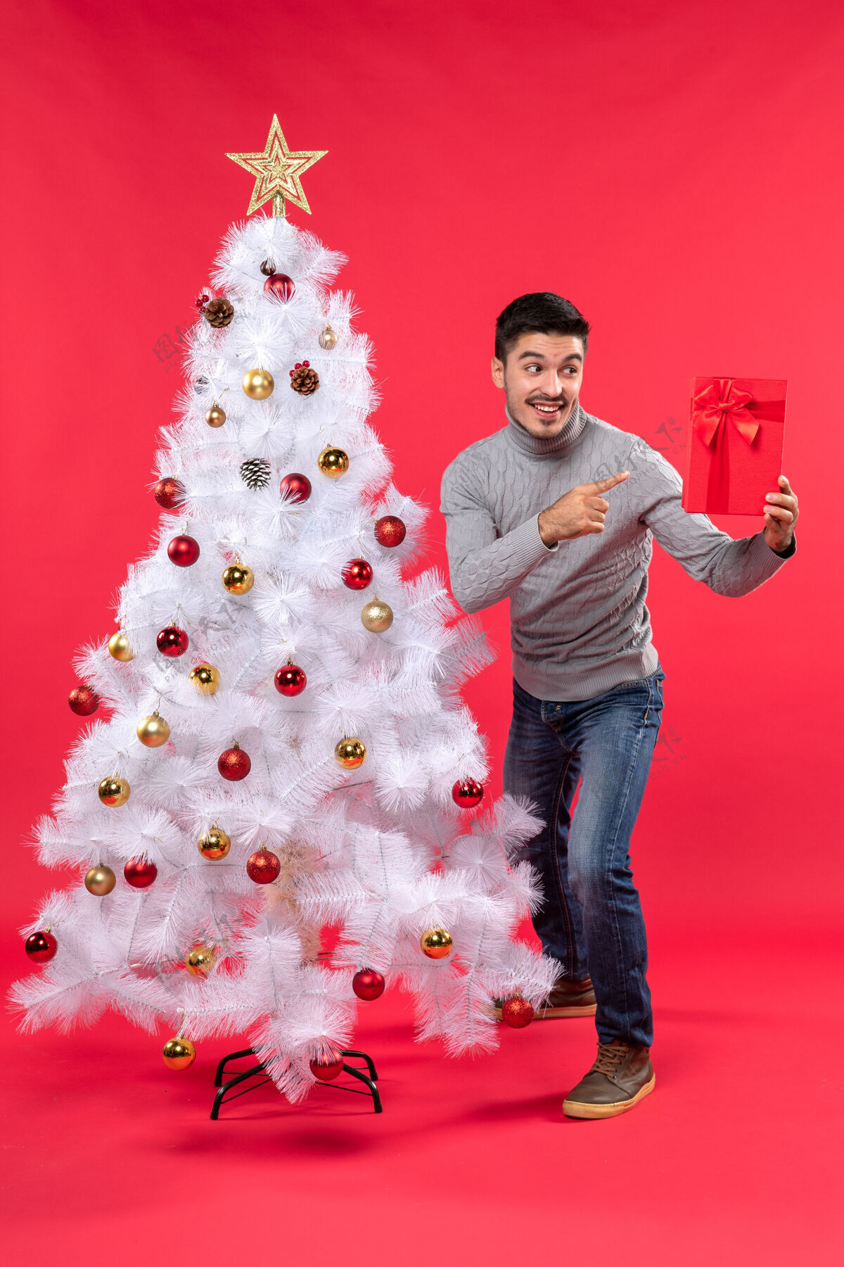 指着俯视图：年轻人站在装饰好的白色新年树旁 手里拿着他的礼物圣诞树庆祝礼物