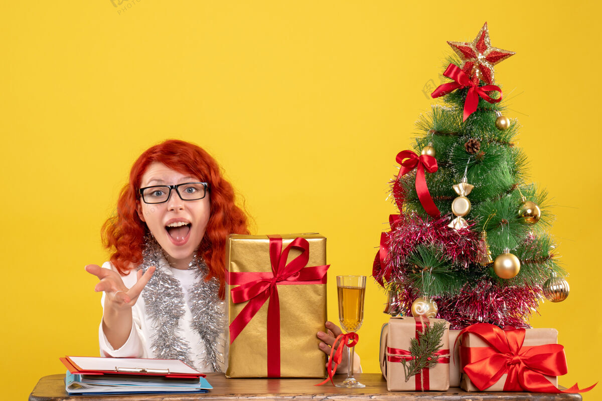 女性前视图：女医生拿着圣诞礼物和黄色背景上的圣诞树坐着礼物圣诞医生