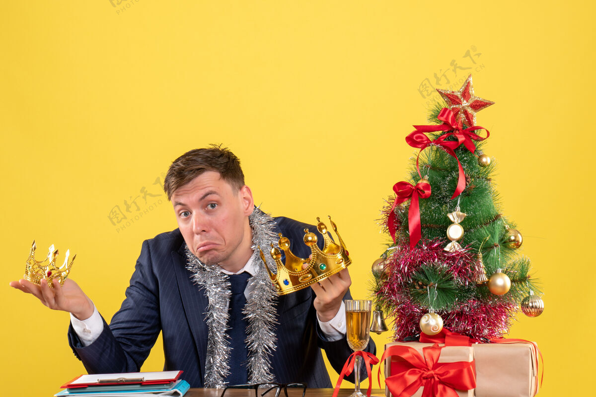 花商务人士双手捧着皇冠坐在圣诞树旁的桌子前 黄色的礼物抱着男人黄色