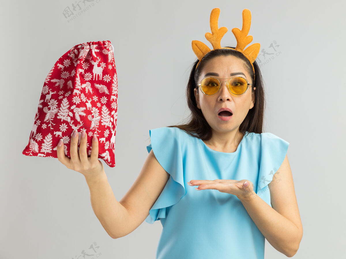 包身穿蓝色上衣的年轻女子 戴着滑稽的鹿角边 戴着黄色眼镜 手里拿着圣诞红包 看上去很惊讶地用手把它呈现出来圣诞女人圣诞快乐