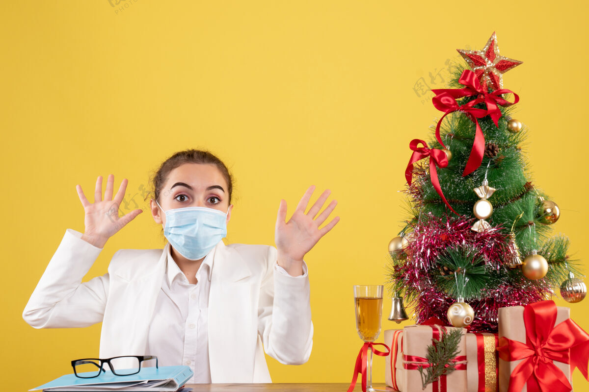 前面正面图女医生戴着防护面具坐在黄色背景上 戴着圣诞树和礼品盒专业防护人
