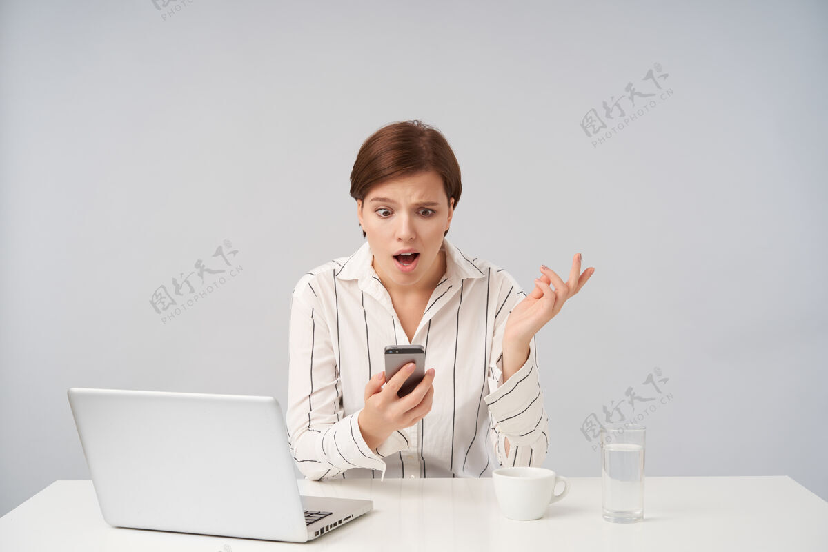 女商人困惑的棕色头发的年轻短发女性手持手机 困惑地看着屏幕 困惑地举起手掌坐在白色的椅子上衣服棕色屏幕