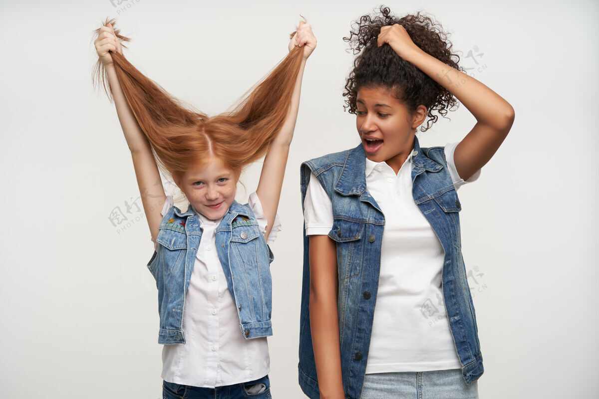 自然室内写真可爱欢快的红发女童子玩她的长发 而站在白色与微笑的黑皮肤黑发女士积极有趣肖像
