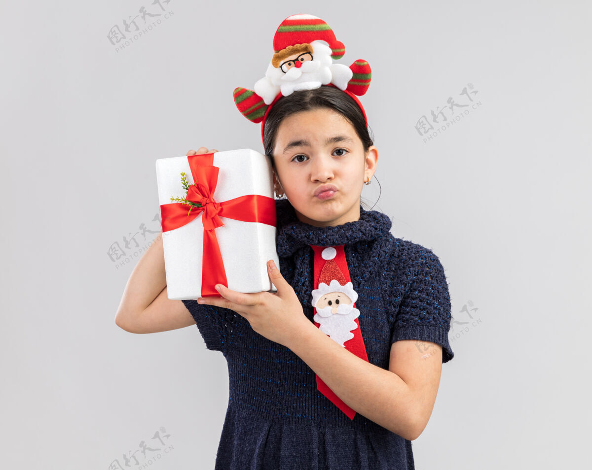 戴着小女孩穿着针织连衣裙 头上系着一条红色领带 头上戴着一个滑稽的圣诞圈 手里拿着圣诞礼物 神情困惑 表情悲伤编织快乐衣服