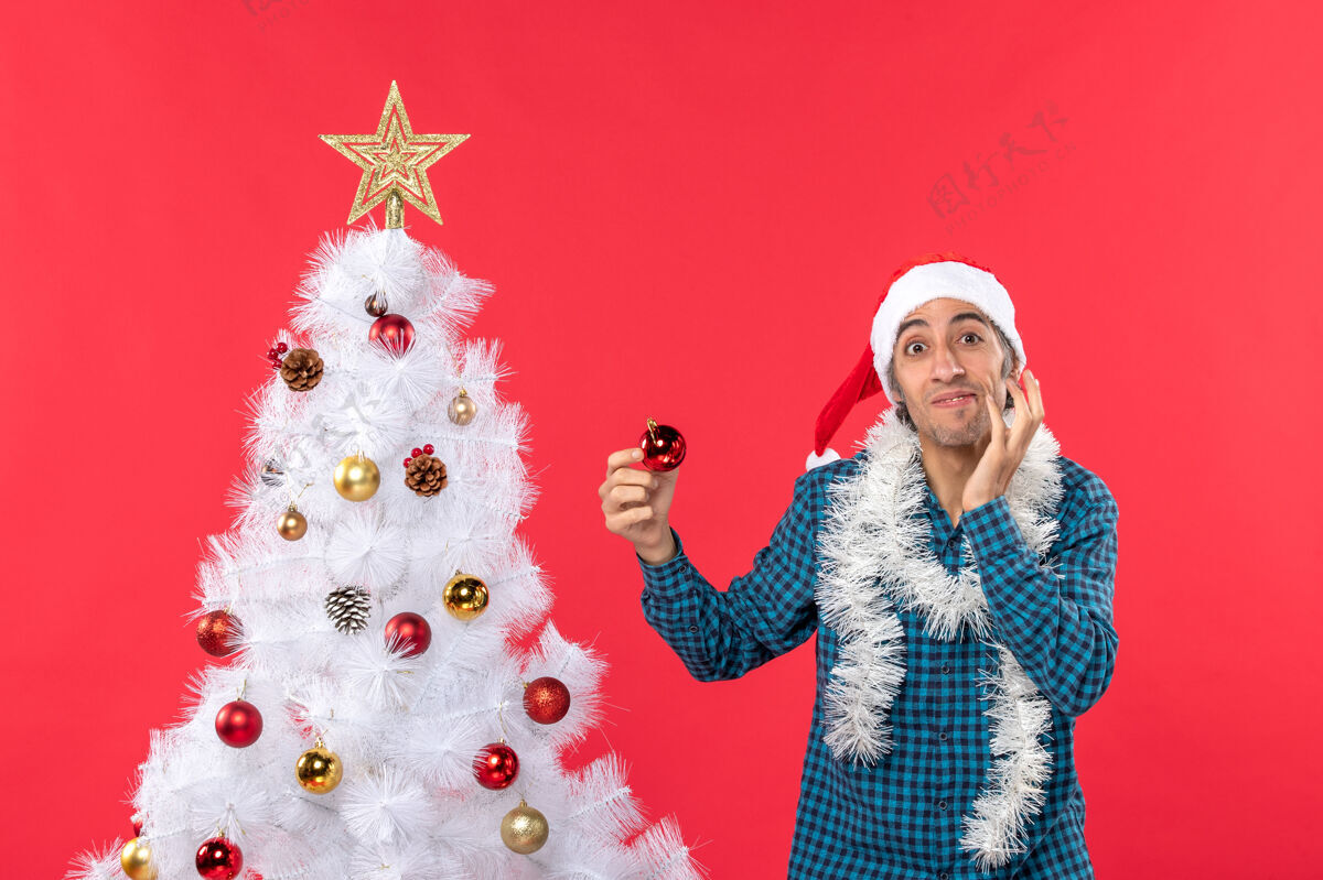 圣诞困惑的年轻人戴着圣诞老人的帽子 穿着蓝色的衬衫 手里拿着装饰品圣诞老人思想帽子