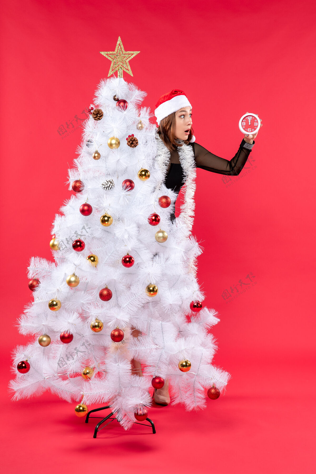 圣诞老人圣诞气氛与穿着黑色连衣裙 戴着圣诞老人帽子的年轻漂亮女士藏在新年树后面时钟雪人衣服
