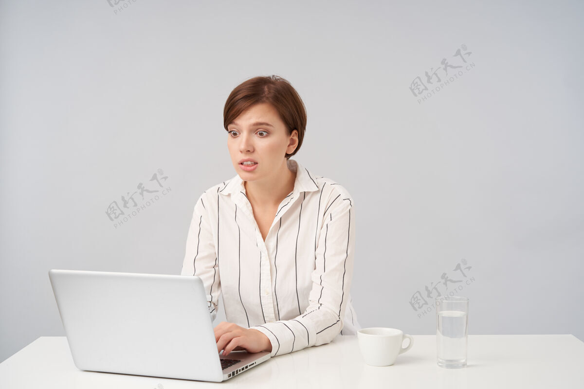 衣服激动的年轻短发黑发女性坐在白色的椅子上 穿着条纹衬衫 一边用笔记本电脑在键盘上打字 一边困惑地看着自己正式办公室肖像