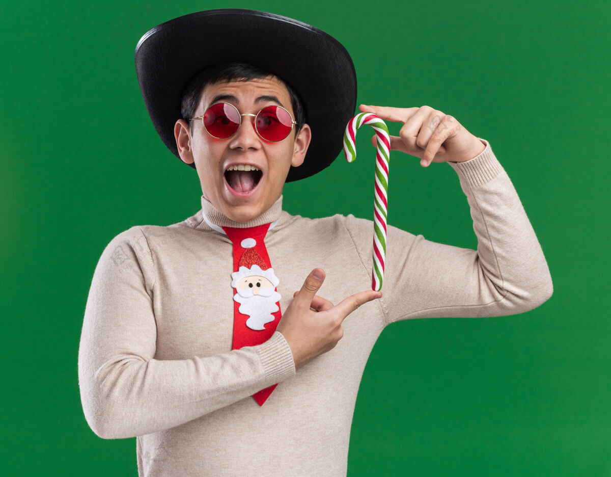 糖果兴奋的年轻人戴着帽子 打着圣诞领带 戴着眼镜 手里拿着圣诞糖果 被隔离在绿色的墙上兴奋年轻帽子