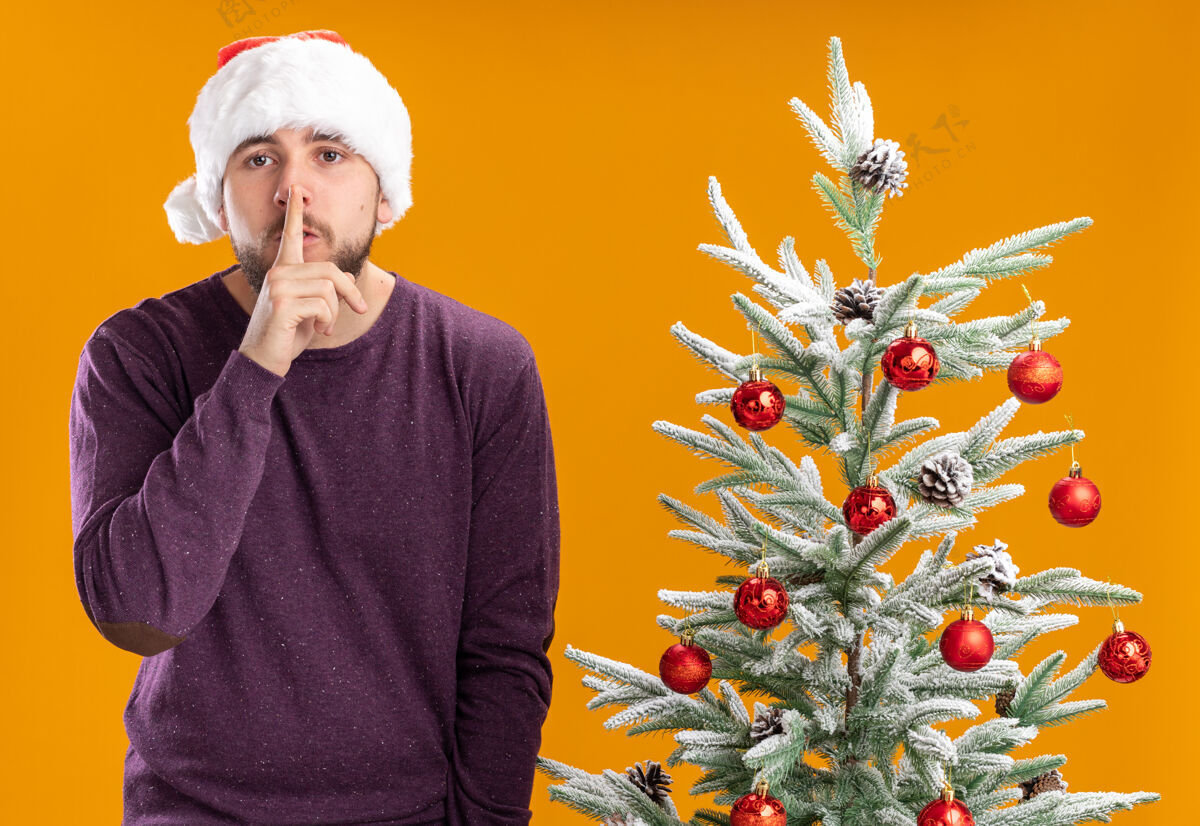圣诞树身穿紫色毛衣 戴着圣诞帽的年轻人看着摄像机在橙色背景下的圣诞树旁做着沉默的手势圣诞看毛衣