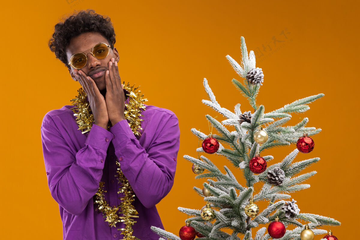 眼镜悲伤的美国黑人年轻人戴着一副戴着金箔花环的眼镜 站在橙色背景的装饰过的圣诞树旁穿着圣诞节金属片