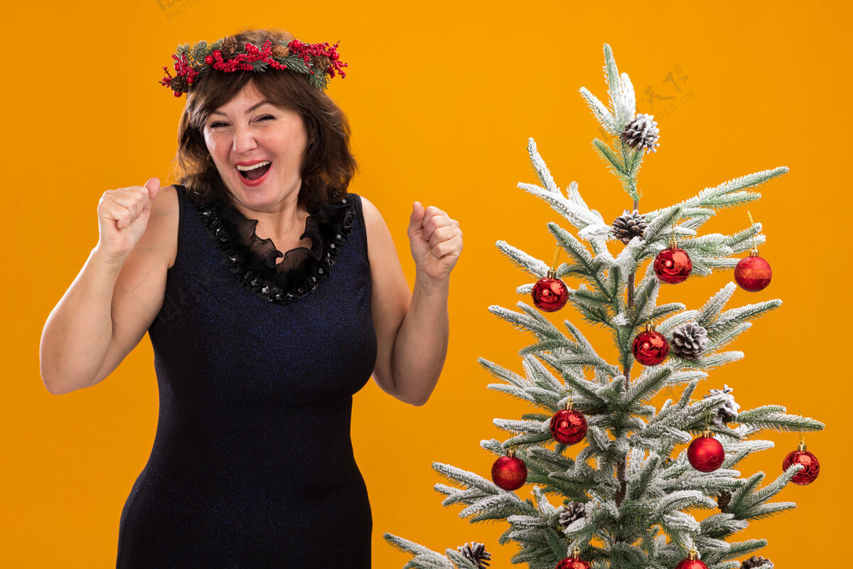 金属片快乐的中年妇女头戴圣诞花环 脖子上戴着金箔花环 站在装饰好的圣诞树旁看花环是的