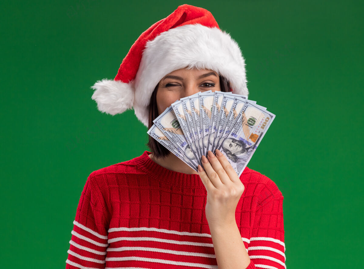圣诞节自信的年轻女孩戴着圣诞帽从后面拿着钱在绿色的墙上眨眼孤立眨眼帽子圣诞老人