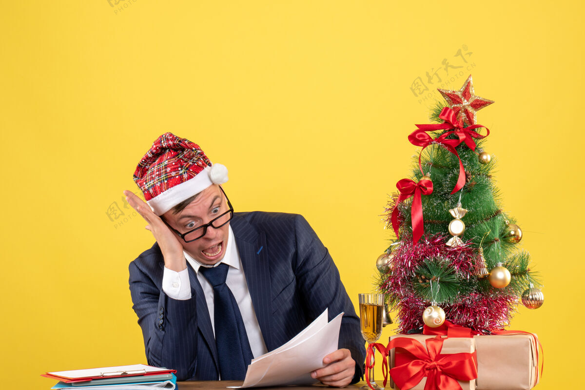 桌子前视图震惊的男子坐在圣诞树附近的桌子上 黄色的礼物男人圣诞节震惊的男人