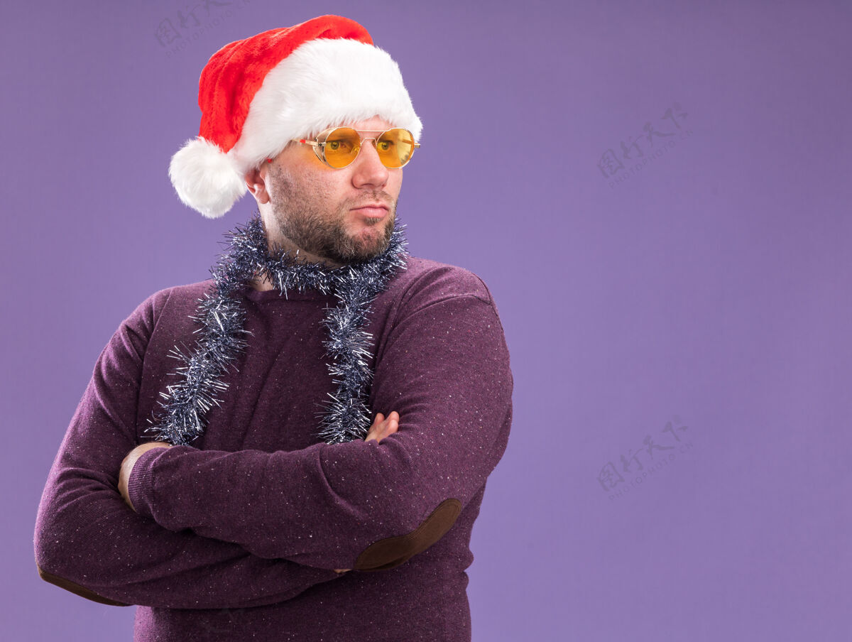 花环自信的中年男子戴着圣诞帽 脖子上戴着金箔花环 戴着眼镜 站着 闭着身子 看着紫色墙壁上孤立的一面 留着复制空间站着圣诞节穿着