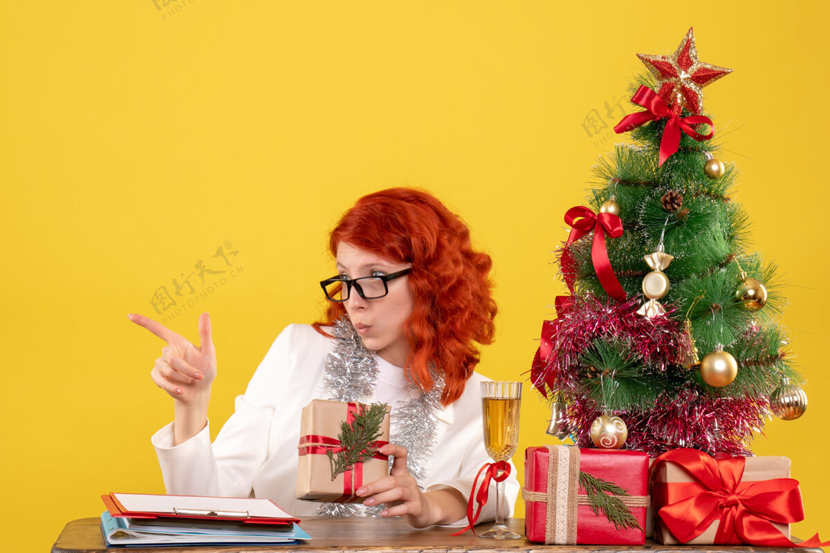 圣诞节前视图女医生坐在桌子后面 黄色背景上有圣诞礼物 还有圣诞树和礼品盒年份微笑女医生