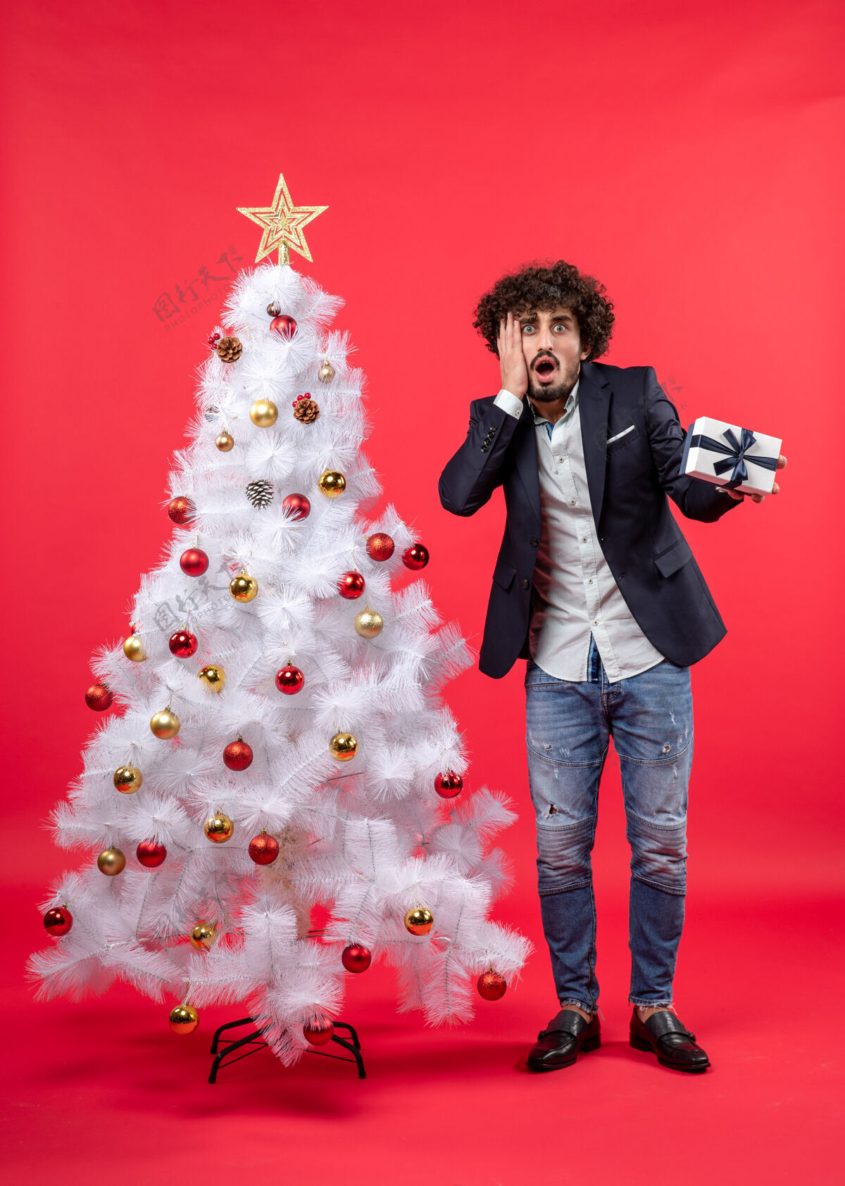年轻人精疲力尽的年轻人拿着他的礼物站在红色的右边装饰的白色圣诞树旁成年人一边年轻人