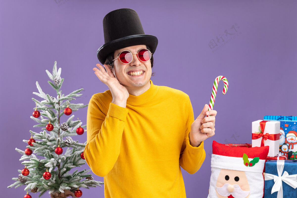快乐快乐的年轻人穿着黄色高领毛衣 戴着黑色帽子 手持糖果手杖 站在一棵圣诞树旁 愉快地微笑着 紫色背景下的礼物年轻人圣诞树穿着