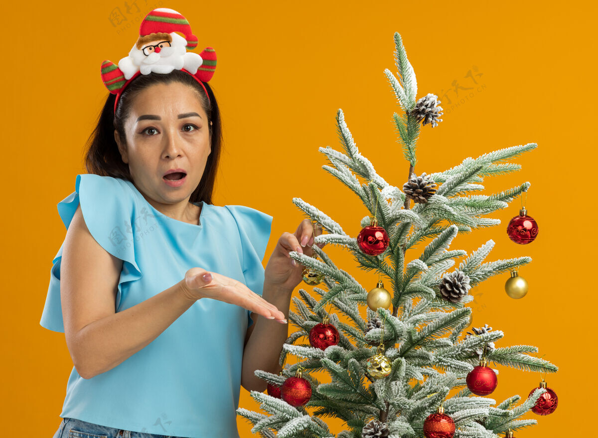 有趣身穿蓝色上衣的年轻女子戴着有趣的圣诞环装饰圣诞树 看起来很困惑 站在橙色背景上不高兴地举起手臂圣诞橙色边缘
