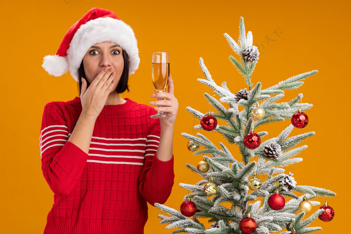 保持一个戴着圣诞帽的年轻女孩站在装饰好的圣诞树旁 手里拿着一杯香槟 手放在橘色的墙上 嘴巴被隔离圣诞节靠近杯子
