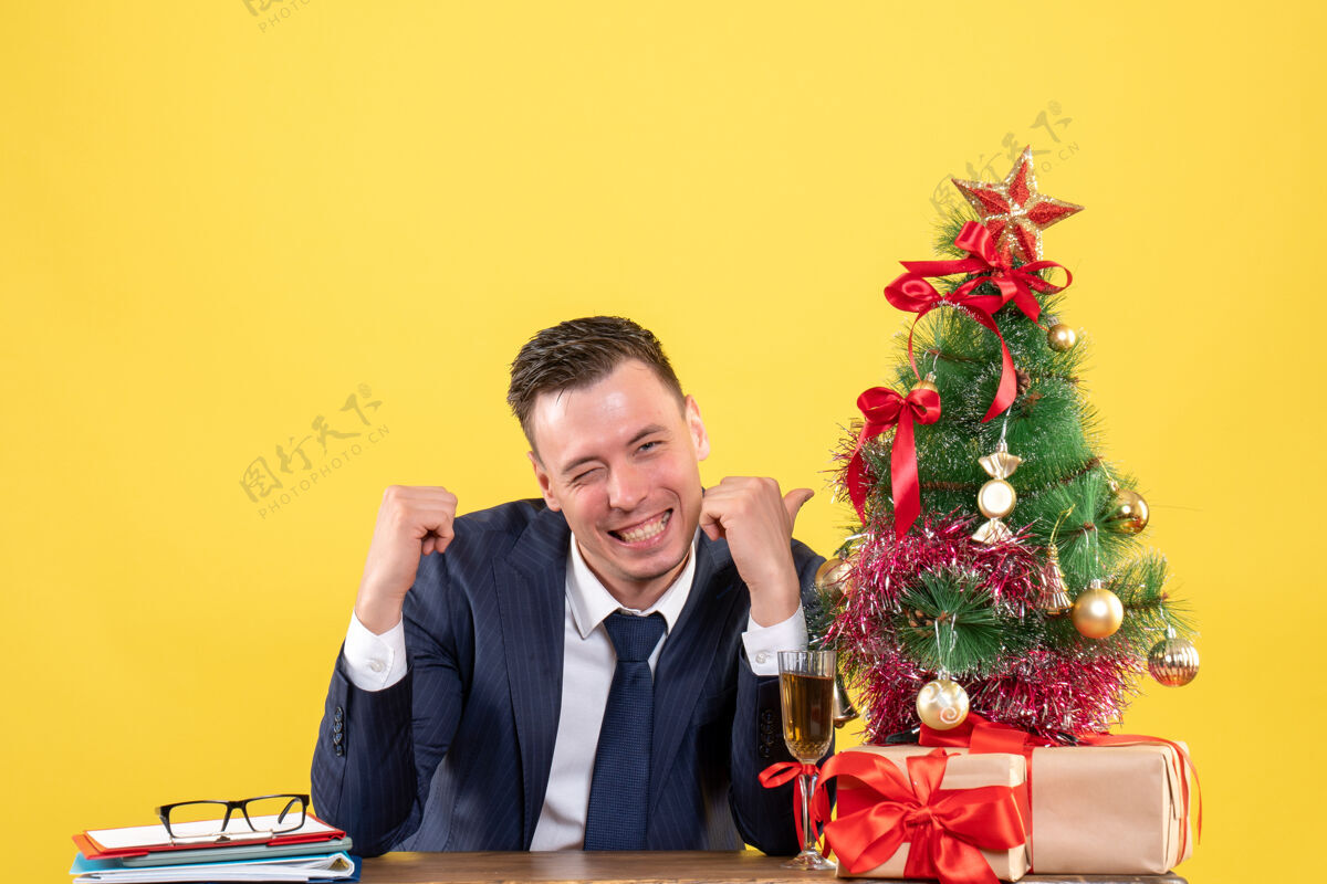 桌子正面图微笑的男子指着圣诞树坐在圣诞树旁边的桌子上 黄色的礼物办公室主管团队