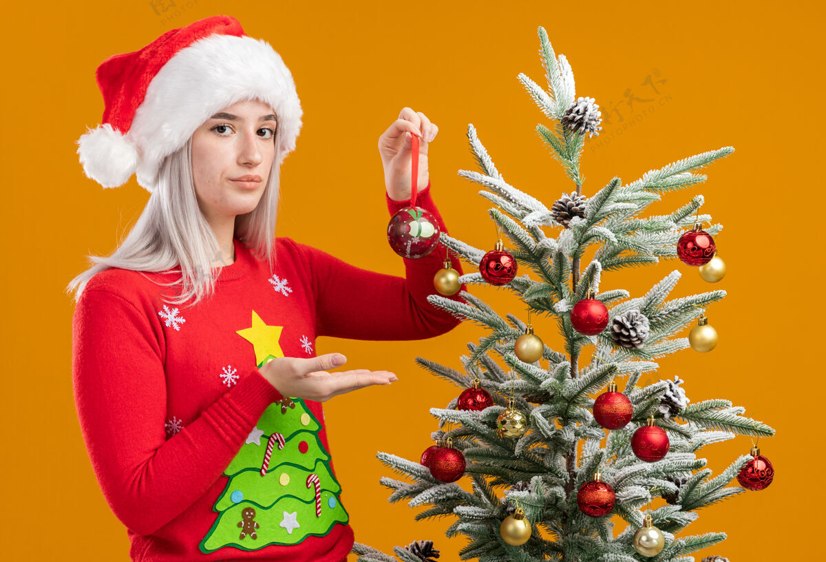 抱着严肃的金发女郎 穿着圣诞毛衣 戴着圣诞帽 手举圣诞球 站在橙色背景下的圣诞树旁圣诞树球帽子
