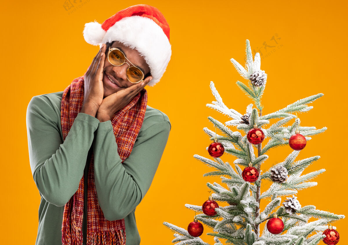 圣诞节戴着圣诞帽 脖子上围着围巾的非裔美国人站在橙色墙上的圣诞树旁 快乐而积极围着正面围巾
