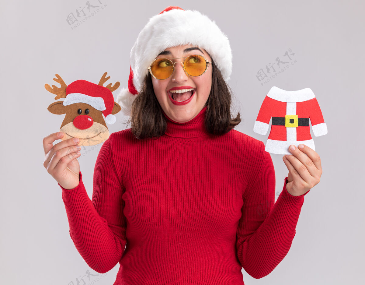 圣诞节快乐的小女孩穿着红色毛衣 戴着圣诞帽 戴着眼镜 拿着圣诞玩具 脸上挂着微笑 站在白色的墙上穿着玩具圣诞老人