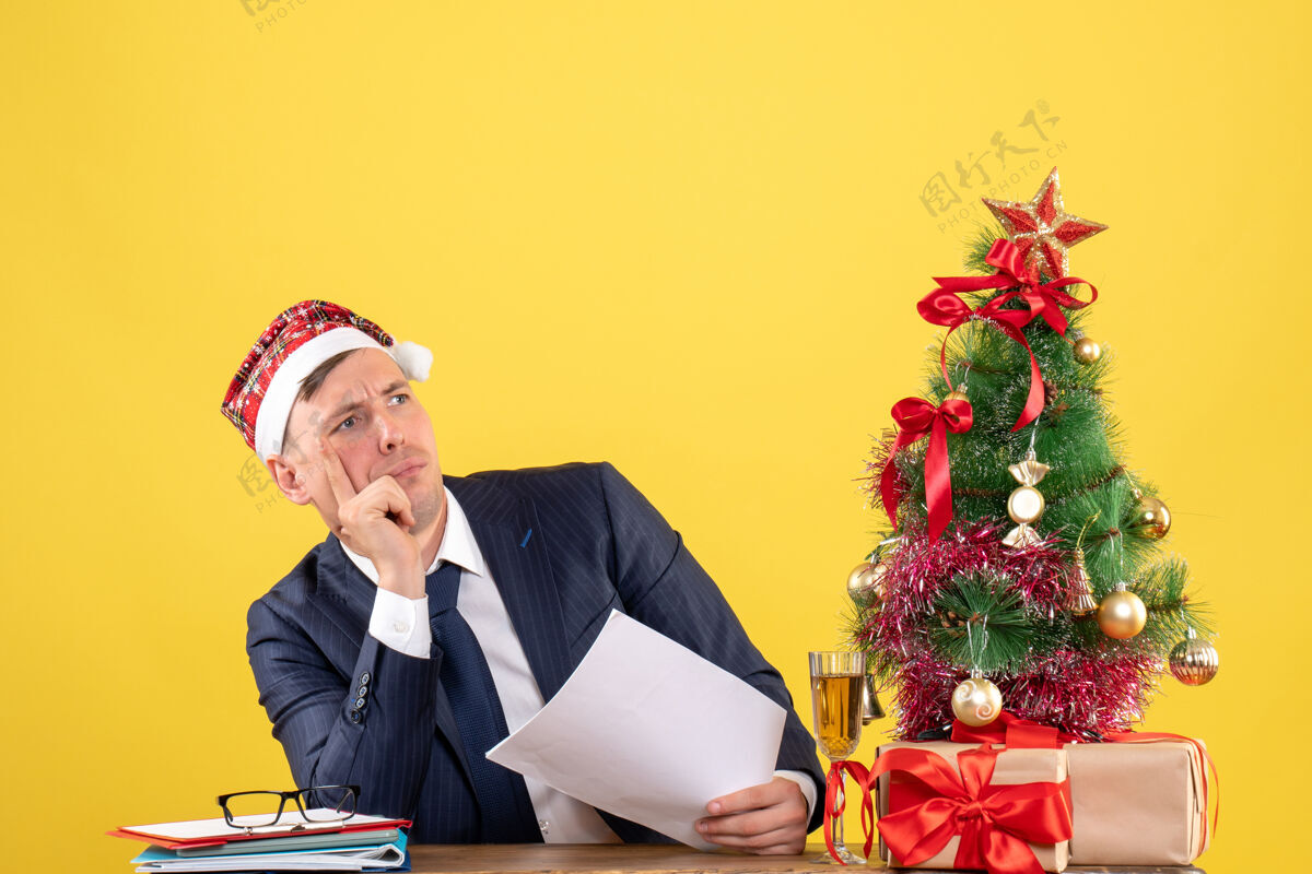 男人一个体贴的男人戴着圣诞帽坐在圣诞树旁的桌子前 黄色的礼物坐着商人圣诞老人