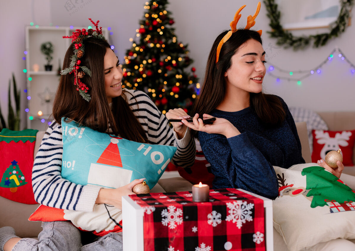圣诞节可爱的年轻女孩们戴着冬青花环和驯鹿头带 手里拿着玻璃球饰品和电话 坐在扶手椅上 在家里享受圣诞节时光坐着扶手椅头带